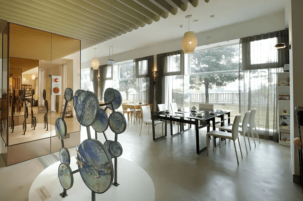 【草屯住宿】草屯知達工藝會館的用餐空間兼展覽廳，中央擺設藝術雕塑作品，玻璃落地窗引入戶外綠意，黑白簡約風桌椅營造藝術感，左側展示空間可見各類藝術品與文創商品，環境靜謐有格調。