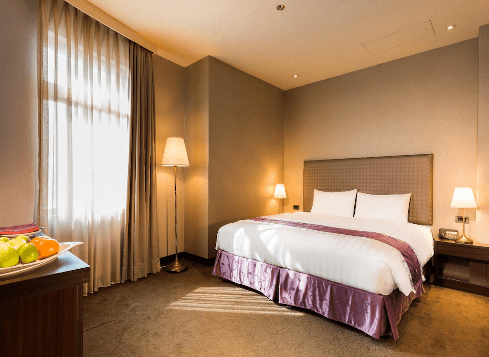 【草屯住宿】草屯福美大飯店的飯店雙人房，配有一張大床、柔和的燈光與落地窗，紫色床裙與床尾巾增添一絲華麗氣息，房內設有邊桌與檯燈，整體設計溫馨舒適。