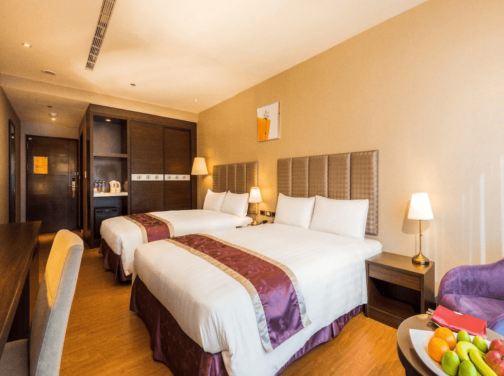 【草屯住宿】草屯福美大飯店的雙人雙床房型，兩張大床併排擺放，配有紫色床巾和床裙，牆上掛有簡約畫作，房內設有書桌、檯燈、沙發椅與迎賓水果，整體空間寬敞明亮。