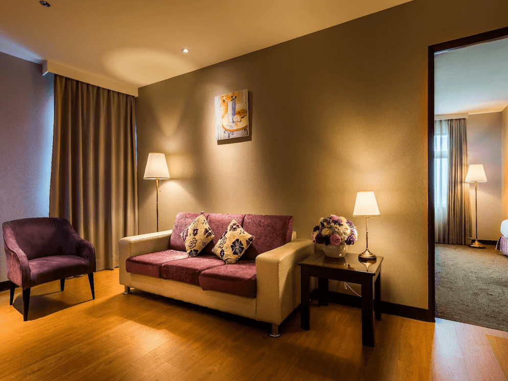 【草屯住宿】草屯福美大飯店的客廳區域，設有一張紫色雙人沙發與單人沙發椅，搭配溫馨的檯燈與落地燈，牆上懸掛著簡約畫作，旁邊可見通往臥室的門口，氛圍靜謐溫馨。