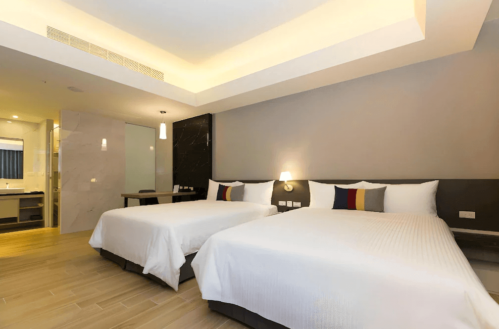 【蘆洲住宿】雀客旅館-新北蘆洲的四人房，配有兩張雙人床，床鋪潔白整齊，牆面為柔和灰色調，天花板設有間接照明。房內有一張工作桌與吊燈，衛浴區採用半開放式設計，整體空間寬敞舒適。