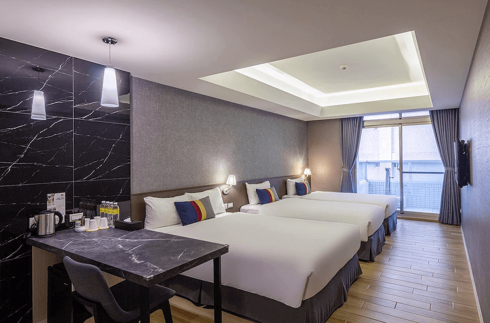 【蘆洲住宿】雀客旅館-新北蘆洲的四人房空間，配有三張標準床位，搭配灰藍色窗簾與現代風格照明。靠牆設有大理石紋理工作桌與備品區，室內自然光充足，適合家庭或團體旅客入住。