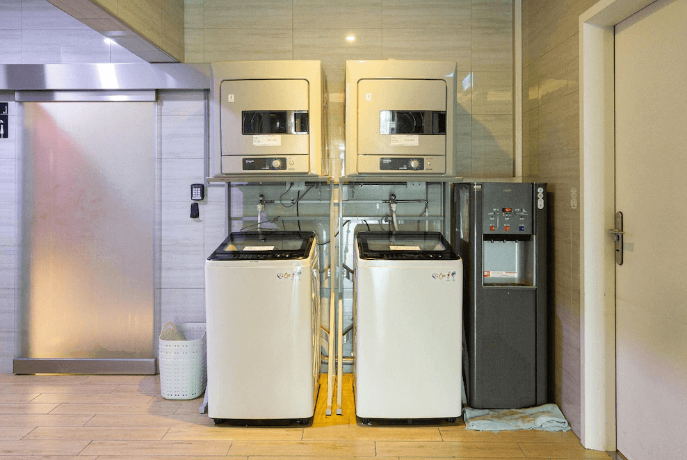 【蘆洲住宿】雀客旅館-新北蘆洲的公用設施區提供兩台洗衣機與兩台烘衣機，旁邊設有飲水機與洗衣籃，方便長住旅客自助洗衣。空間整潔明亮，牆面與地板採用淺木紋設計。