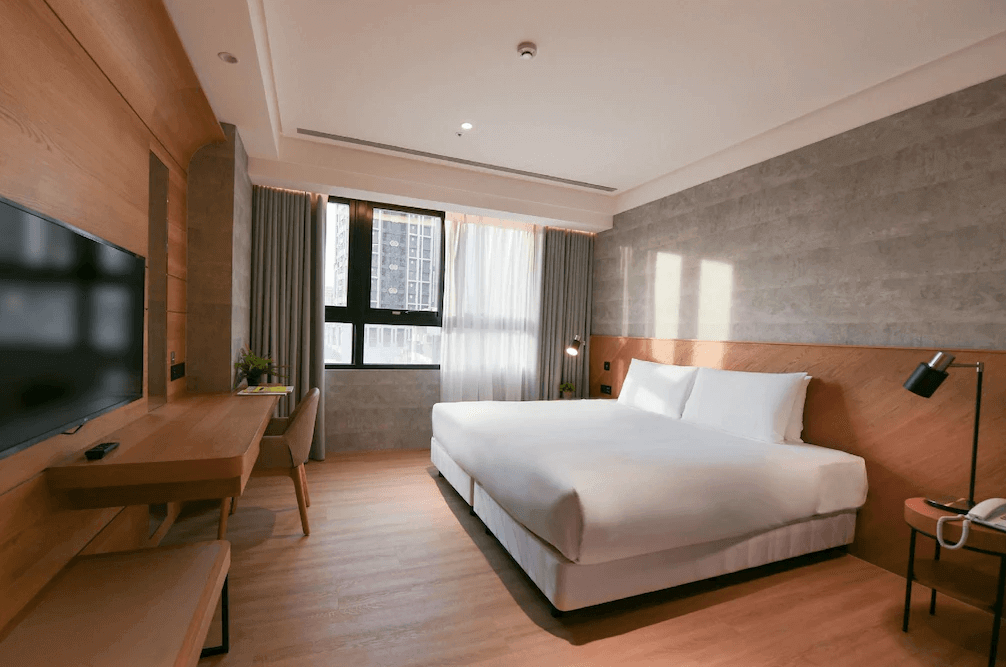 【三重住宿】雀客藏居 - 新北三重水漾的大床房，融合灰色與木質元素，營造簡潔質感氛圍。窗邊擺放書桌與觀景椅，採光良好，房內設有壁掛電視與床邊閱讀燈，功能齊備。