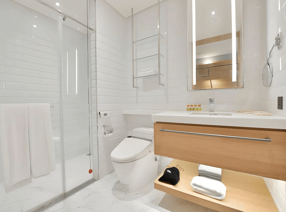 【三重住宿】雀客藏居 - 新北三重水漾的明亮浴室，牆面鋪設白色磁磚，配備智慧馬桶、玻璃淋浴間與大片鏡面。洗手台下方設有收納空間，整體整潔且現代感十足。