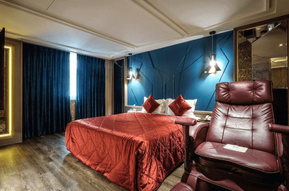 【蘆洲住宿】香奈爾汽車旅館的雙人房，床舖鋪著酒紅色絲絨床罩與靠枕，牆面採用藍色立體幾何線條設計，搭配兩盞吊燈營造溫馨氛圍。房內配有一張深紅色全功能按摩椅，地板為深色木質，整體風格典雅現代。