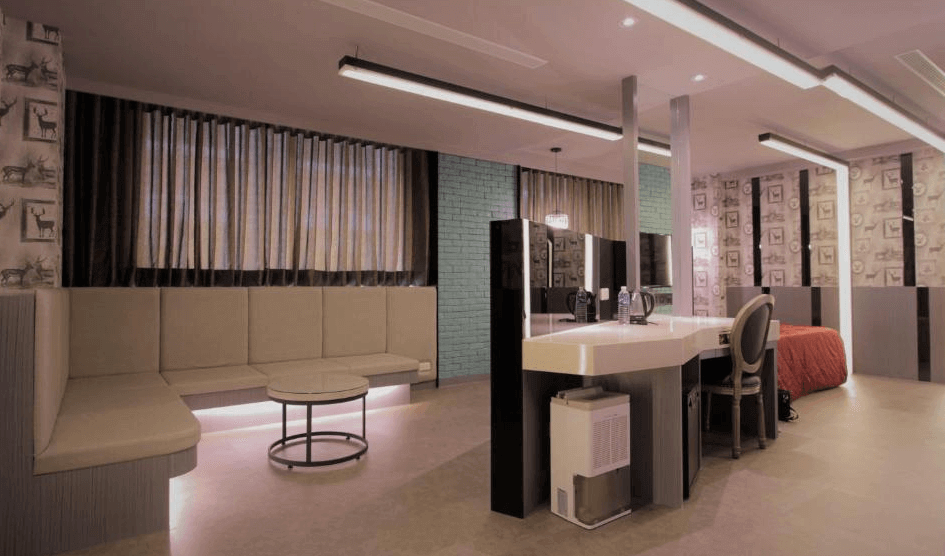 【蘆洲住宿】香奈爾汽車旅館的多功能空間，左側為長型沙發與圓形茶几，中央為化妝台與雙面鏡子，後方是雙人床與設計感強烈的牆面。整體色調為灰白與紅色搭配，展現現代與個性風格。