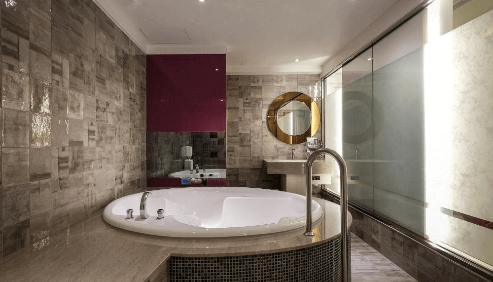 【蘆洲住宿】香奈爾汽車旅館的豪華衛浴空間，牆面與地板鋪設灰色磁磚，中央為圓形按摩浴缸，後方設有洗手台與金色圓鏡。右側為大片玻璃隔間，引入自然光線，營造明亮舒適的沐浴環境。