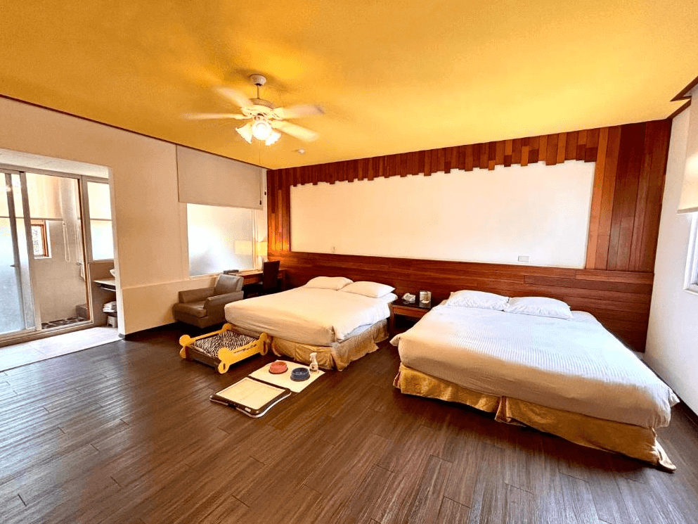 【楊梅住宿】麗多森林溫泉酒店的四人房，設有兩張雙人床，木質地板與牆面搭配黃色天花板，營造溫馨氛圍。房內備有寵物床、飼料碗與尿布墊，適合攜帶毛孩入住，左側設有獨立陽台空間與沙發休憩區。