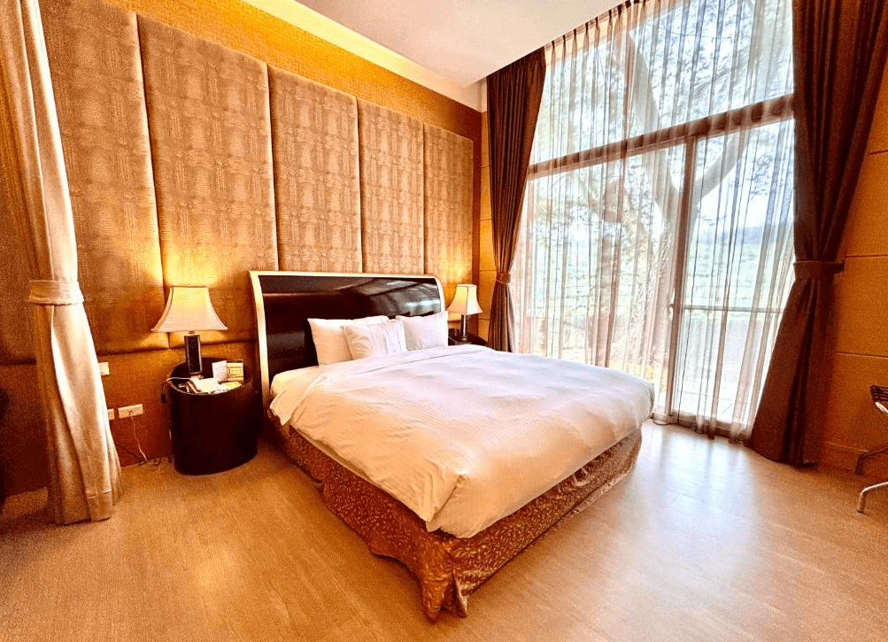 【楊梅住宿】麗多森林溫泉酒店的高挑落地窗套房，設有一張舒適雙人床，金棕色牆面與絨布床頭板營造典雅氛圍。窗簾半掀，可見窗外綠意盎然的森林與山景，自然光灑入室內，環境明亮清新。