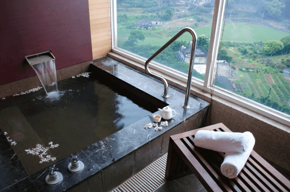 【楊梅住宿】麗多森林溫泉酒店的室內景觀湯屋，設有石造泡湯池，窗邊為大片景觀玻璃，可眺望遠方田園風光與山景。池畔放有茶壺、茶杯與毛巾，營造放鬆泡湯氛圍，水面漂浮著裝飾花瓣，環境清幽雅緻。