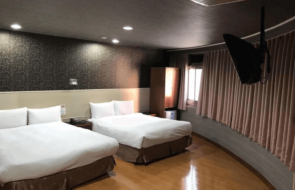 【左營住宿】龍翔大飯店的黑色花紋牆面與暖色燈光營造溫馨氛圍，房內有兩張大床、木質地板及弧形牆面設計，附有壁掛式電視。