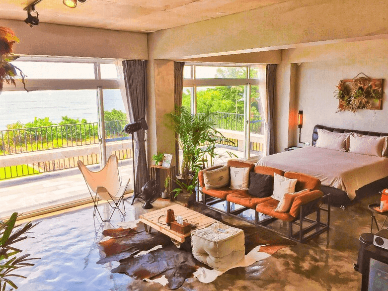 【豐濱住宿】Noosa 海岸行館的另一間海景套房，房內配置舒適雙人床、復古風格沙發與木製茶几，落地窗外可欣賞綠意與海景，整體風格融合自然與工業元素，打造放鬆愜意的度假空間。