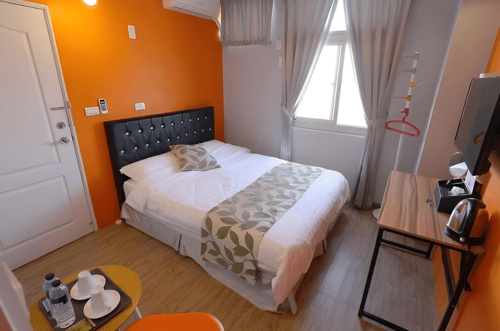 【布袋住宿】光影民宿溫馨雙人房，空間雖小但設備齊全，搭配鮮明橘色牆面與典雅灰色窗簾，附有電視、熱水壺與小型書桌，適合單人或情侶短住。