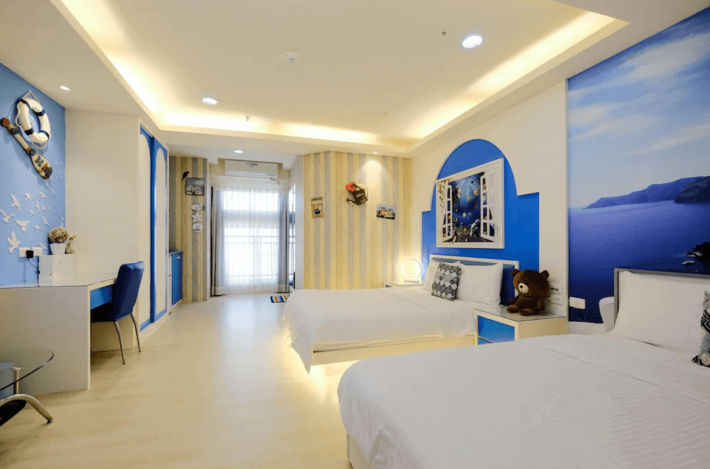【高雄寵物友善住宿】八五亞太旅店的房內設計以藍白為主色調，牆面有地中海風格窗景與海景壁畫，配置兩張雙人床與書桌，整體空間寬敞明亮，充滿童趣與渡假氛圍，適合家庭旅遊入住。