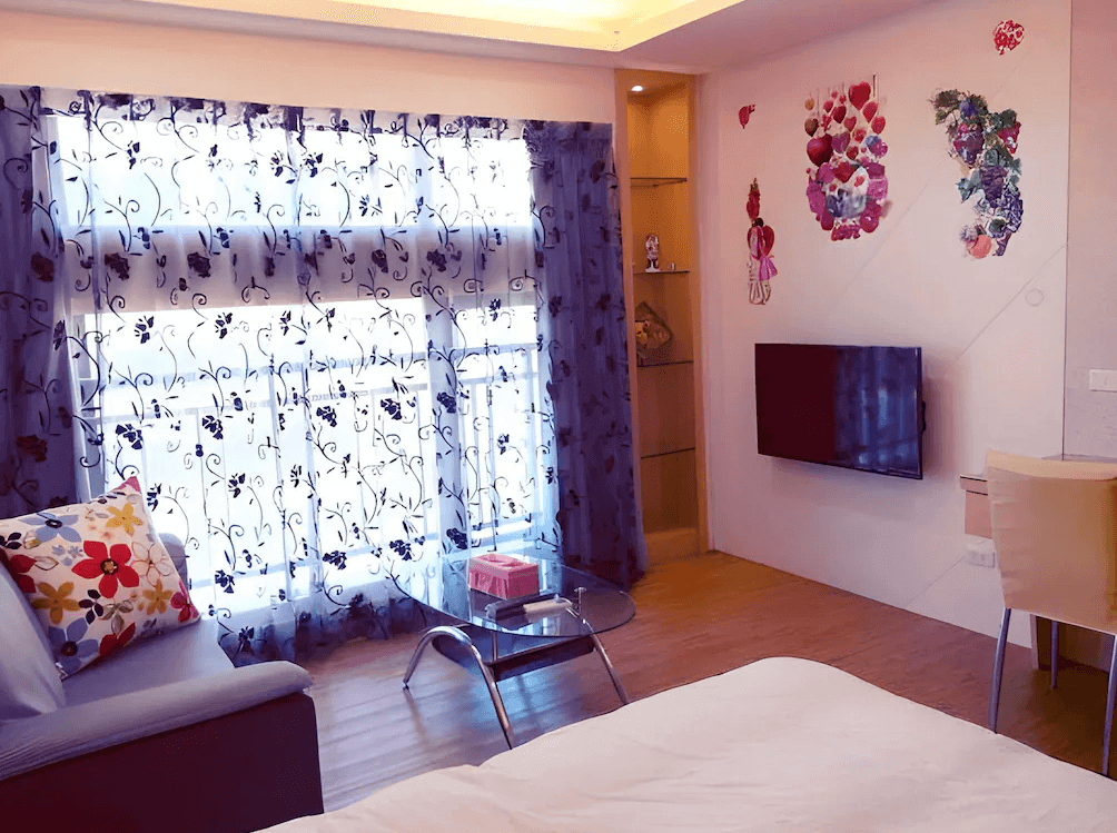 【高雄寵物友善住宿】八五亞太旅店的房內佈置以粉色與花卉圖案為主，窗簾採用黑白花草圖騰，電視牆與玻璃展示櫃裝飾有愛心與可愛貼圖，適合喜愛浪漫與溫馨氛圍的旅客。