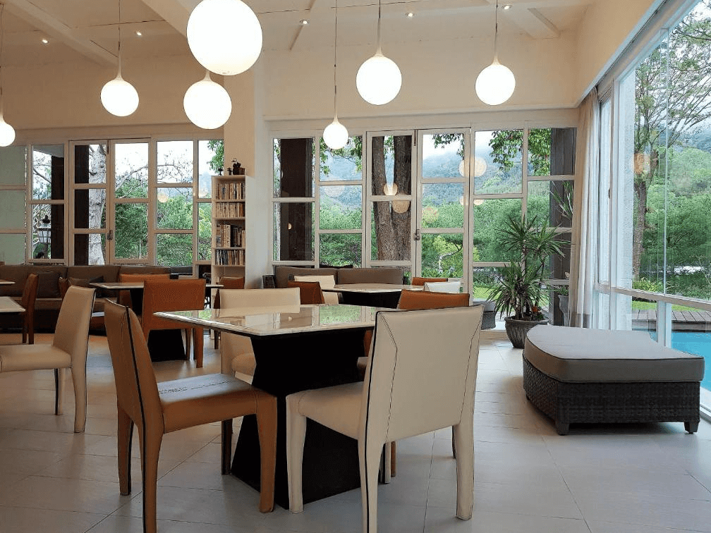 【苗栗包棟民宿】八角隱士莊園的大廳，空間挑高明亮，搭配玻璃落地窗與圓形吊燈，提供舒適沙發與餐桌椅，適合閱讀、聚會與用餐，窗外可眺望山林與泳池景致，營造靜謐自然的休憩氛圍。