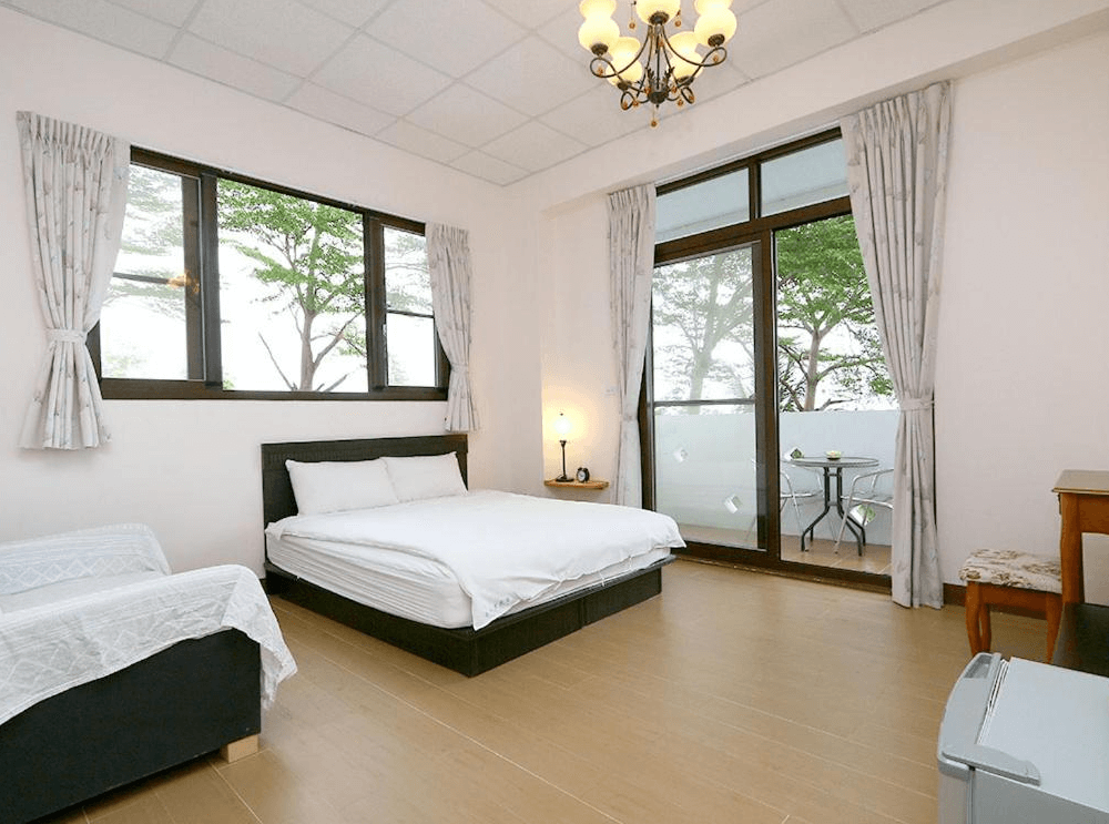 【東河住宿】南八里民宿雙人房，房內設有一張雙人床與沙發椅，大片窗戶與陽台讓室內採光極佳，窗外可見綠樹環繞，空間寬敞簡潔，散發溫馨寧靜的鄉村氛圍。