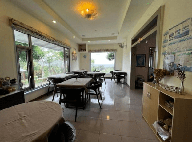 【東河住宿】南八里民宿用餐區，大片窗景可眺望戶外庭園與遠方山海，室內擺設多張方桌與座椅，牆面裝飾旅遊資訊與手作擺飾，提供溫馨又明亮的用餐空間。