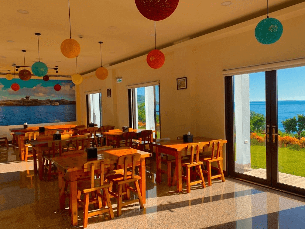 【台東成功住宿】台東「嵐海民宿」餐廳空間，採用木質桌椅搭配鮮豔活潑的吊燈裝飾，大片落地窗外即為湛藍的海洋景色，空間明亮且氣氛愜意，適合悠閒用餐及觀賞海景。