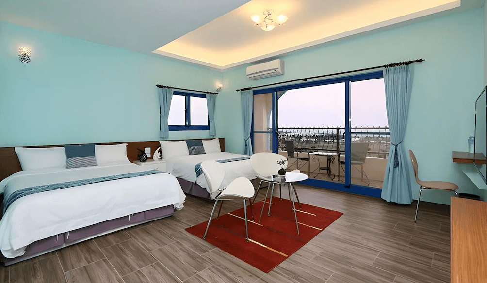 【台東成功住宿】台東「幸福小築民宿」四人房內景，牆面採溫柔的湖水藍色，房內配置兩張雙人床，搭配簡約白色家具與落地窗陽台，整體空間明亮寬敞、風格清新。
