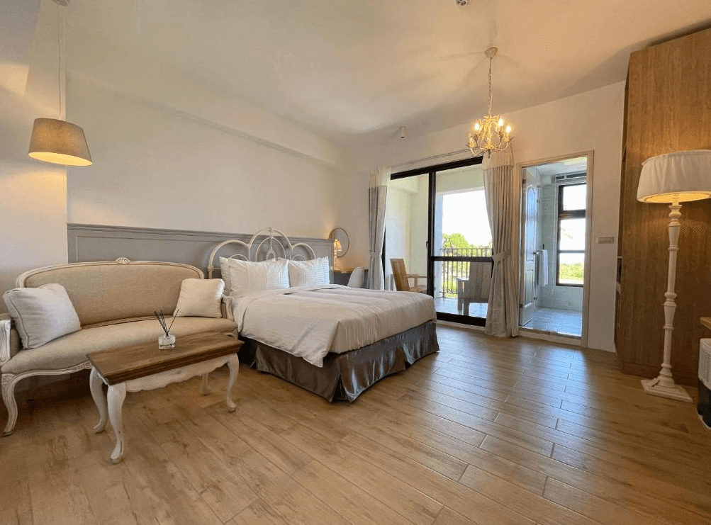 【台東成功住宿】台東海景民宿「奧麗雅安莊園」的雙人房內景，房間設計典雅舒適，配備寬敞的雙人床、精緻沙發座椅組、溫馨燈飾以及可通往戶外陽台的大型玻璃門，整體風格明亮溫暖。