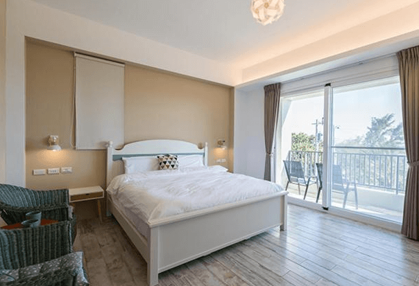 【東河住宿】吉院子民宿典雅雙人房，設有一張白色床架的大床，牆面為溫暖米色系，木質地板營造溫馨氛圍，大片落地窗連接陽台，戶外可見樹景與自然光灑入。