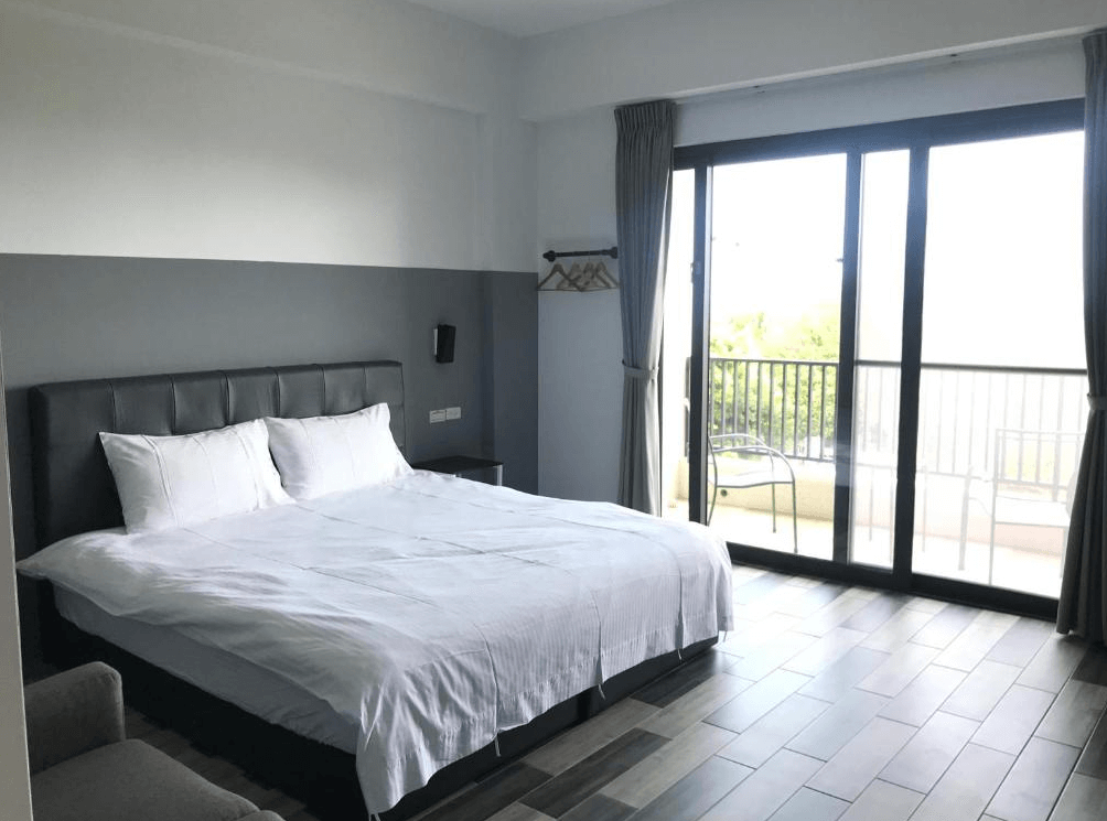 【東河住宿】吉院子民宿現代風格雙人房，搭配深灰色床頭與兩側床頭櫃，房內採用灰白配色牆面，落地窗通往陽台，陽光明亮、視野開闊，簡約舒適。