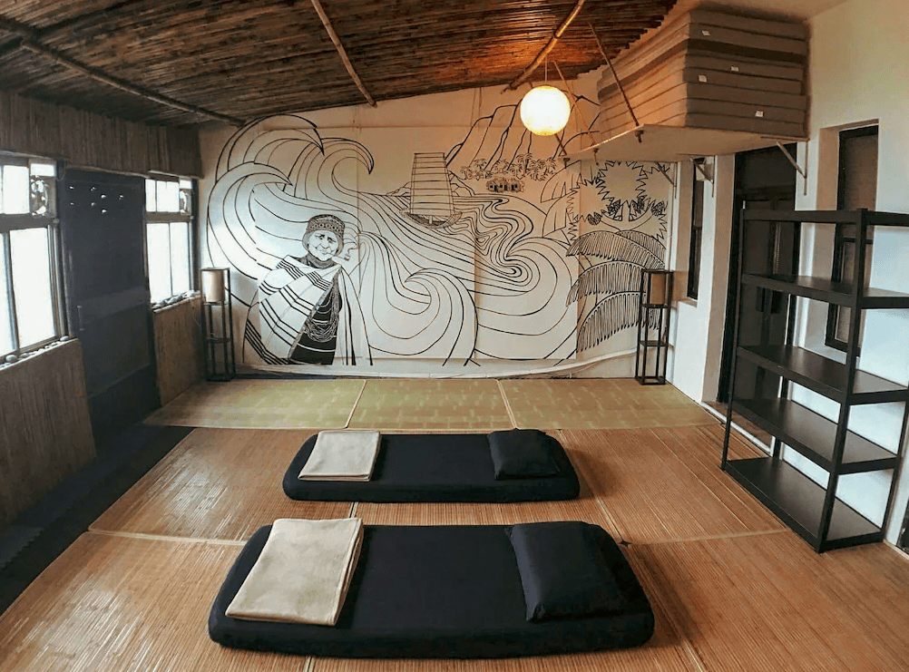 【東河住宿】哇軋力共日式通鋪房，空間鋪設竹蓆與草蓆，設有兩組黑色床墊與毯子，牆上以原住民肖像與山海圖騰繪成手繪壁畫，空間富有自然與文化氛圍，天花板以竹編結構營造出質樸氣息。