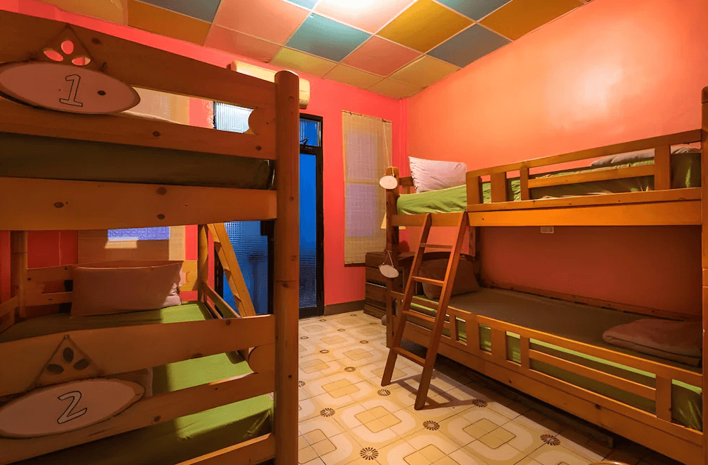 【東河住宿】哇軋力共多人上下舖房，橘色牆面搭配四張木製上下舖，床鋪以綠色床單與柔和燈光點綴，地板為復古磁磚，整體色彩鮮明，充滿活力與溫馨感。