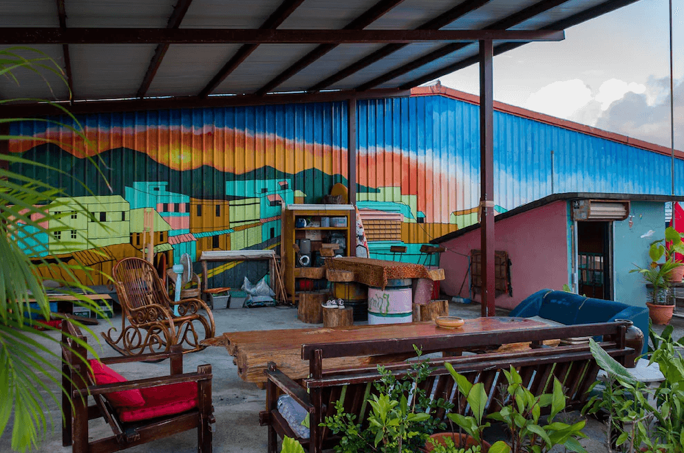 【東河住宿】哇軋力共戶外交誼空間，位於屋頂平台，牆面彩繪熱情繽紛的港口風景，搭配木質桌椅與鞦韆椅，周圍綠意點綴，是放鬆、欣賞日落與與旅人交流的好地方。