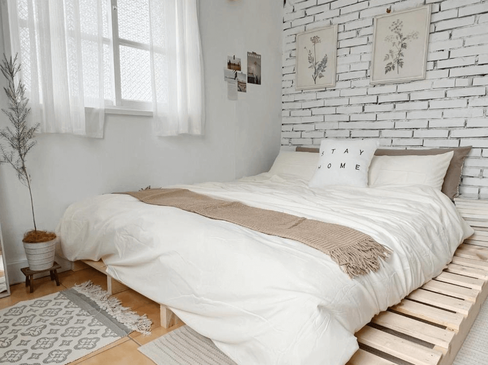 【玉井住宿】山蒔旁白的自然風雙人房，以白磚牆搭配木棧床架展現極簡美感，純白床組搭配米色編織毯與「STAY HOME」抱枕，整體風格溫潤柔和，窗邊垂掛白色紗簾，光線柔和灑落，呈現療癒空間感。