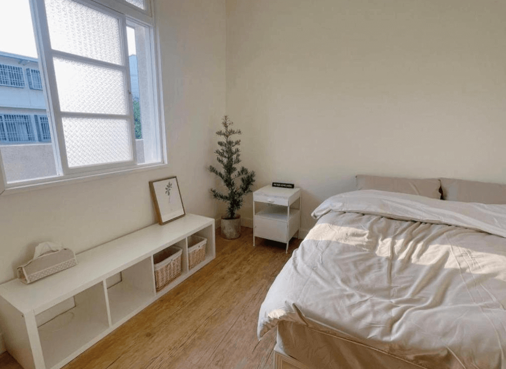 【玉井住宿】山蒔旁白的極簡雙人房，採用淺米與木質色調為主，床鋪柔軟舒適，旁邊搭配白色收納櫃與綠色植栽，窗外為社區街景，自然光線灑落，空間明亮溫馨，給人一種居家放鬆的感覺。