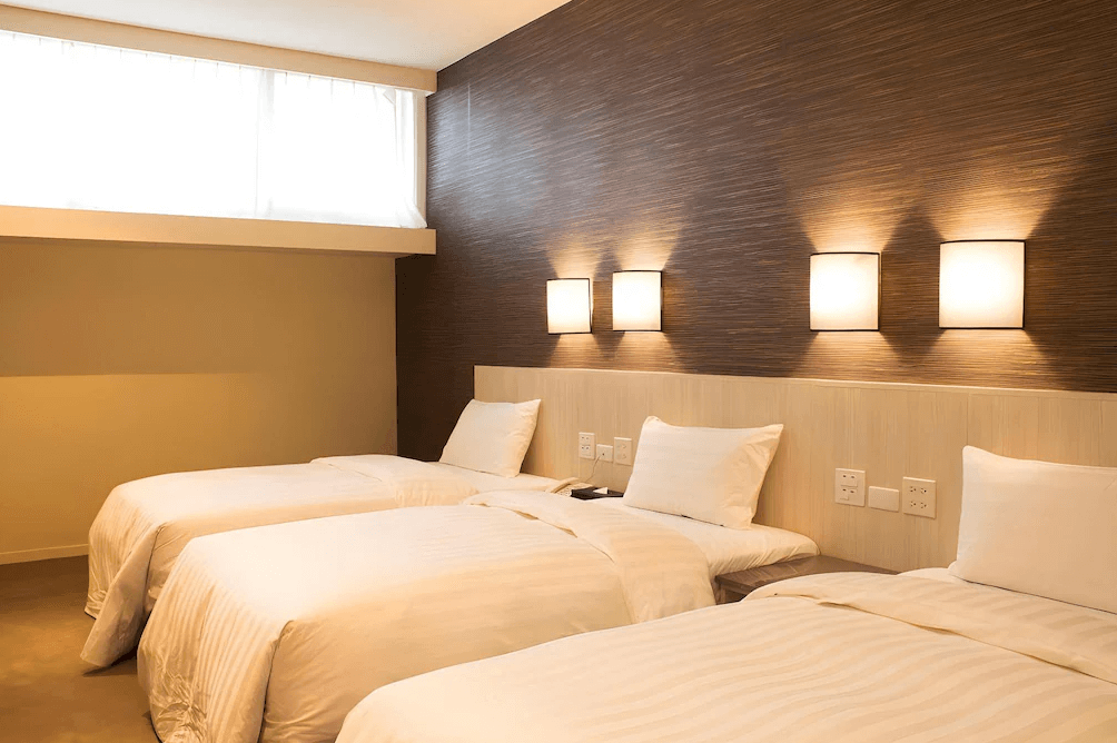 【布袋住宿】布袋文創HOTEL三人房，配有三張單人床，牆面採用溫暖木紋設計，搭配柔和壁燈營造溫馨氛圍，適合家庭或朋友同行入住。
