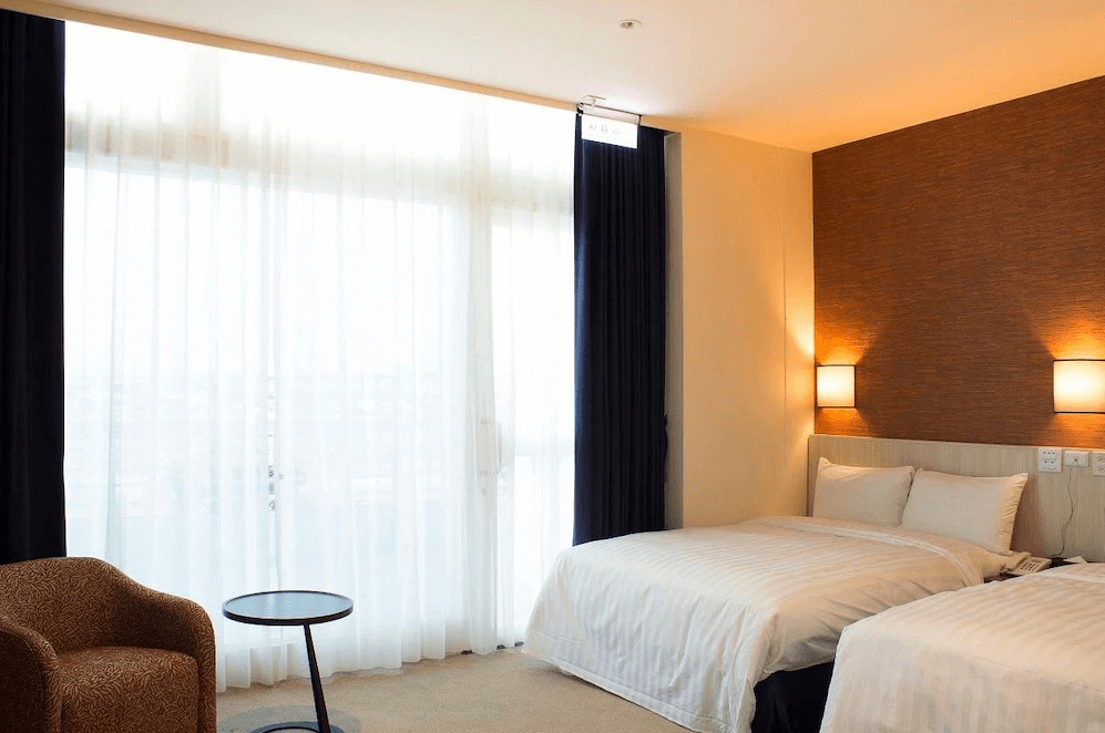 【布袋住宿】布袋文創HOTEL標準雙人房，房內設有兩張單人床，大片白色窗簾搭配深色窗簾，營造寧靜舒適的住宿空間，附設單人沙發與圓形小茶几。