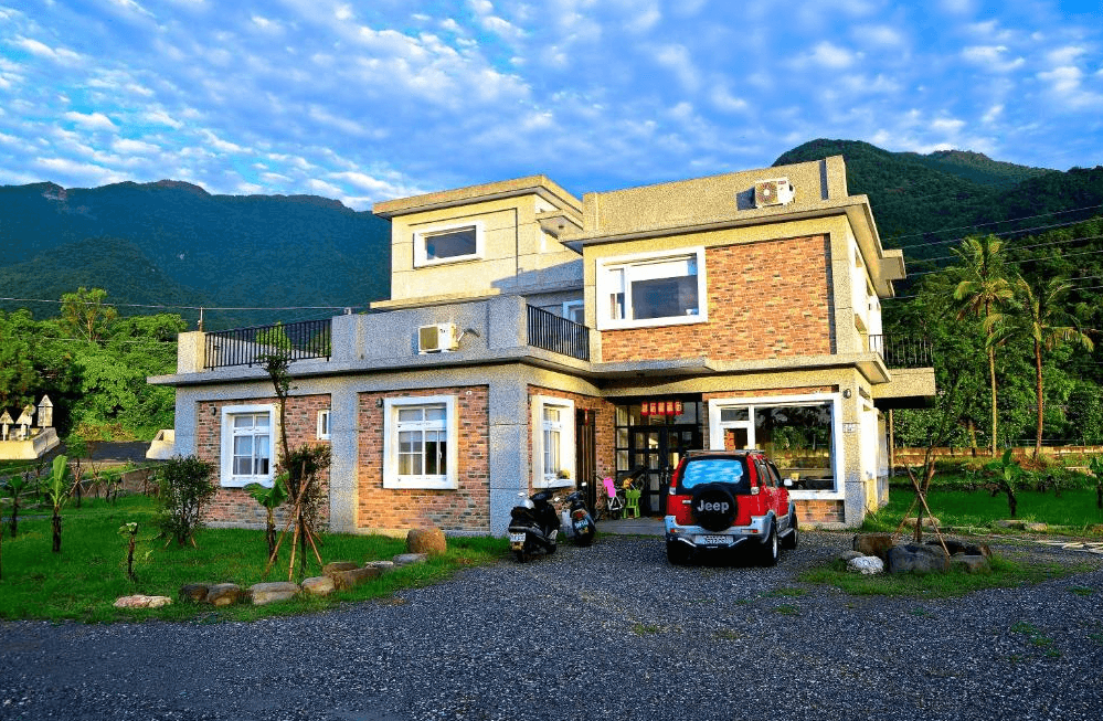 【關山住宿】弋式風莊園民宿的建築外觀，採現代鄉村風格設計，磚牆與灰白色外牆相互映襯，四周環繞著山景與草地，前方停放紅色吉普車與摩托車。