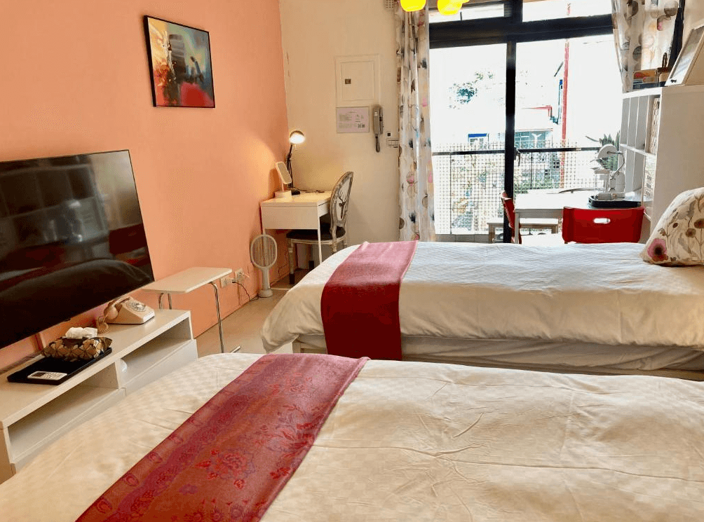 【東河住宿】心適都蘭雙床房型，設有兩張單人床搭配紅色床尾巾與花卉抱枕，牆面為溫暖粉橘色，房內有電視、書桌、落地窗及陽台，自然光充足，整體風格明亮溫馨。