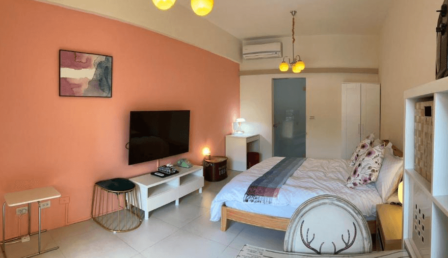 【東河住宿】心適都蘭雙人房，牆面為溫柔粉橘色，配有一張雙人床與花卉枕頭，房內設有電視、小書桌、衣櫃與暖色吊燈，簡約中帶有設計感，空間寬敞舒適。