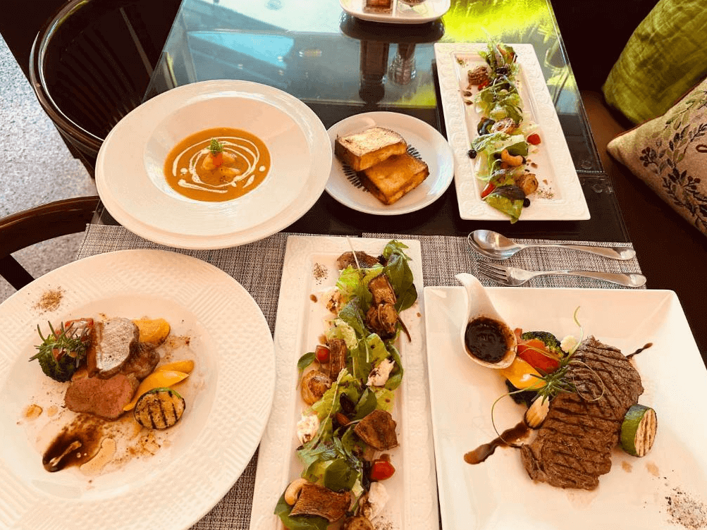 【玉井住宿】恩典之家民宿提供的西式排餐餐點，擺盤精緻多樣，包含牛排、烤蔬菜、濃湯、麵包與沙拉拼盤，搭配透明玻璃桌與自然採光，營造出優雅放鬆的用餐氛圍。