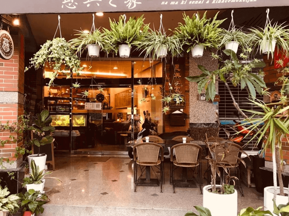 【玉井住宿】恩典之家民宿的門口外觀，玄關以滿佈綠意的垂吊植物點綴，入口兩側擺放多種盆栽，戶外設有休憩座椅，玻璃門後方可見溫暖燈光與餐廳內部，展現出熱情迎賓的氣氛。