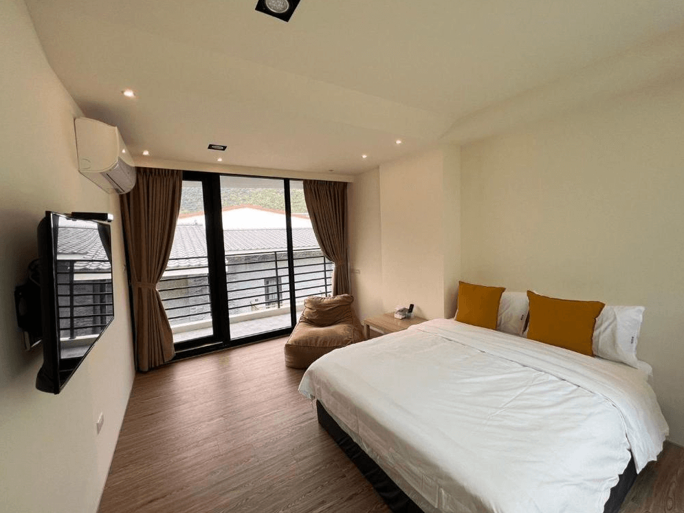 【關山住宿】懶得棟民宿的雙人房，配置簡約舒適的雙人床與芥黃色抱枕，大片落地窗外可見山景與屋頂，室內光線明亮、氛圍溫馨。