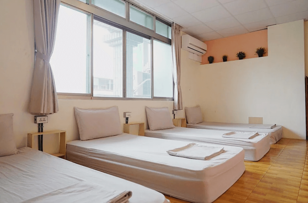 【東河住宿】旅行蟲背包客棧的四人榻榻米房型，設有四張簡約床墊與白色寢具，窗邊採光良好，牆面與地板為木質與米色系搭配，空間清爽溫和，適合團體或朋友入住。