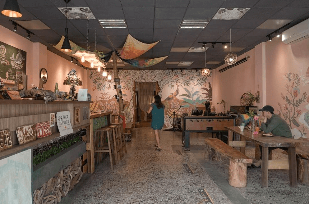 【東河住宿】旅行蟲背包客棧大廳交誼區，牆面繪有熱帶植物插畫，空間內擺放長桌、吧台與桌上足球台，提供旅客交流與放鬆的多元空間，整體氛圍活潑而有趣。