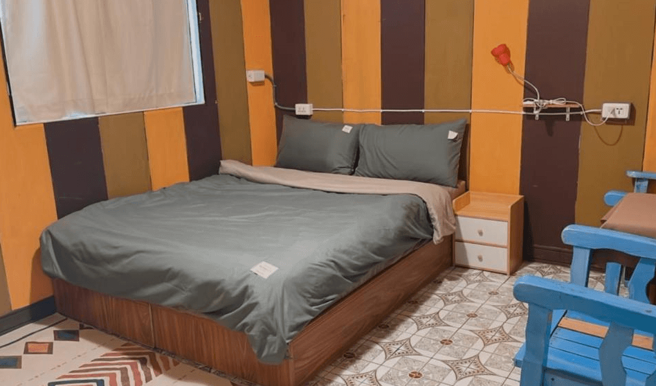 【東河住宿】晃輕鬆客棧咖啡館的雙人房，房間以暖色調條紋牆面裝飾，房內設有舒適的雙人床、床頭櫃以及藍色木質椅子，空間布置簡潔溫馨。