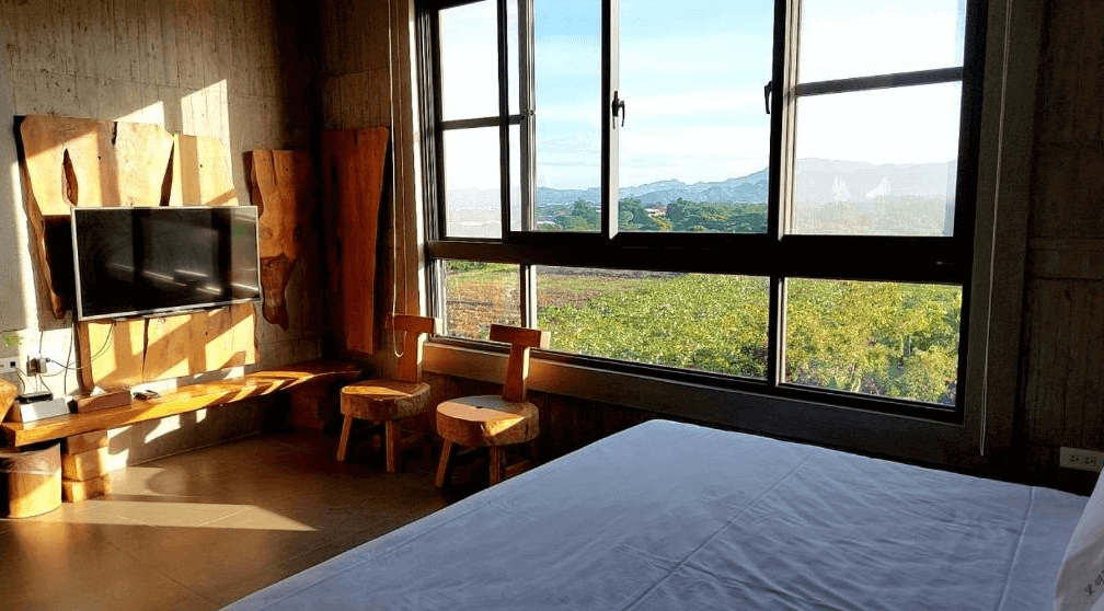 【玉井住宿】望明79民宿房間窗邊視角，床鋪旁可見大片開窗與窗景，向外望去即是綠意田野與遠方山脈，房內電視牆與座椅皆以原木打造，營造自然質樸的入住體驗。