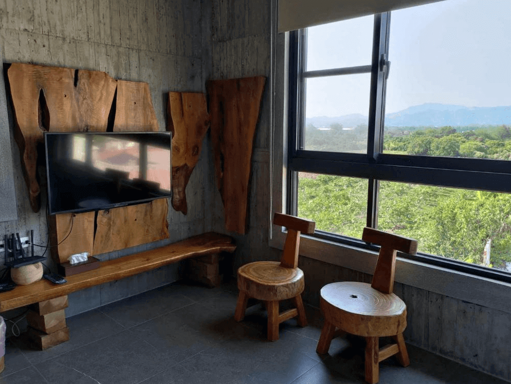 【玉井住宿】望明79民宿客房內的木質電視牆與窗景座位區，電視牆面以不規則實木板設計，搭配手作風格的原木椅與小圓桌，窗外可俯瞰大片綠意田園與遠山景致，質感與自然兼具。