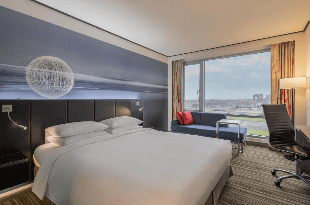 【桃園機場住宿】桃園國際機場凱悅酒店的標準大床房，床頭有藍色系藝術牆面裝飾，房內配有藍色沙發與大面窗戶，可遠眺機場與都市景致。