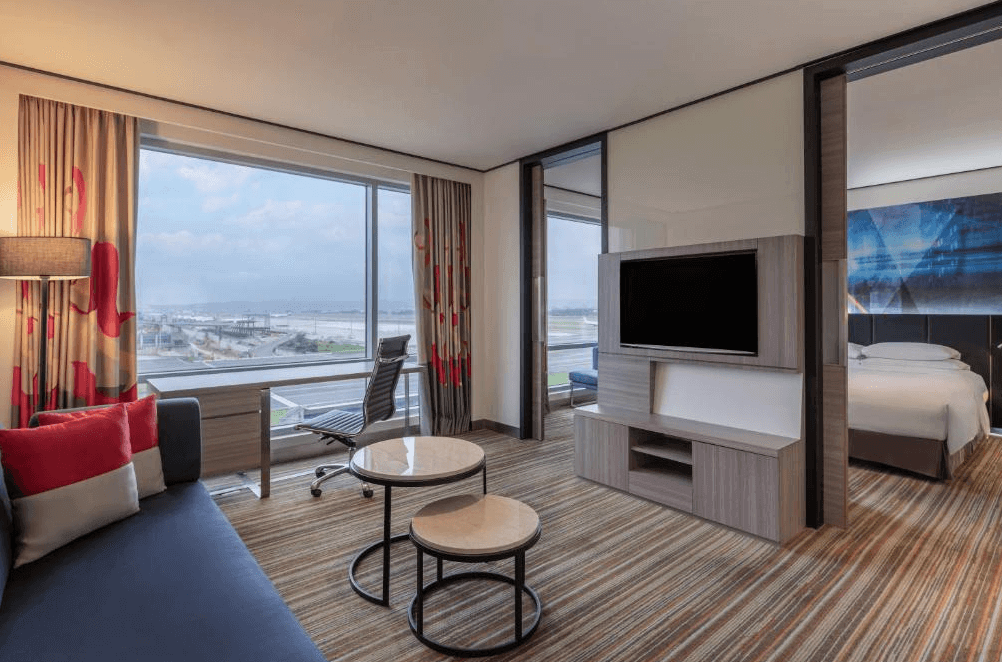 【桃園機場住宿】桃園國際機場凱悅酒店的套房客廳區，設有沙發、書桌與電視牆，連通式設計可通往臥室，窗外為桃園機場跑道與遠景。