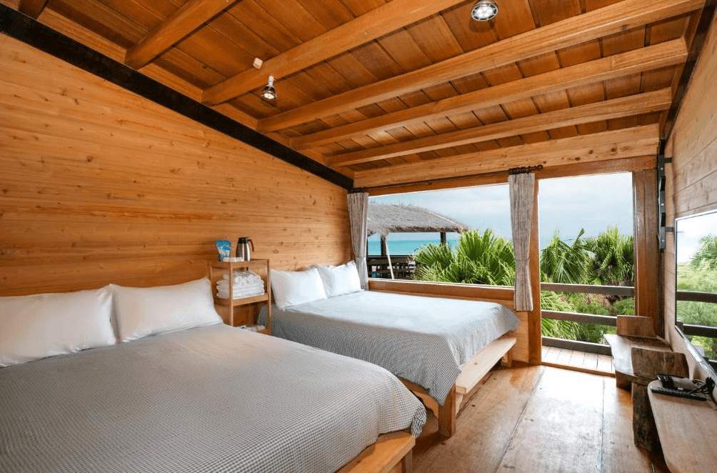 【台東成功住宿】台東「比西里岸營地渡假小木屋」四人房內景，整體採原木建築風格，房內配置兩張大床，大片窗景可直望海景與熱帶椰林，營造出自然愜意的度假氛圍。