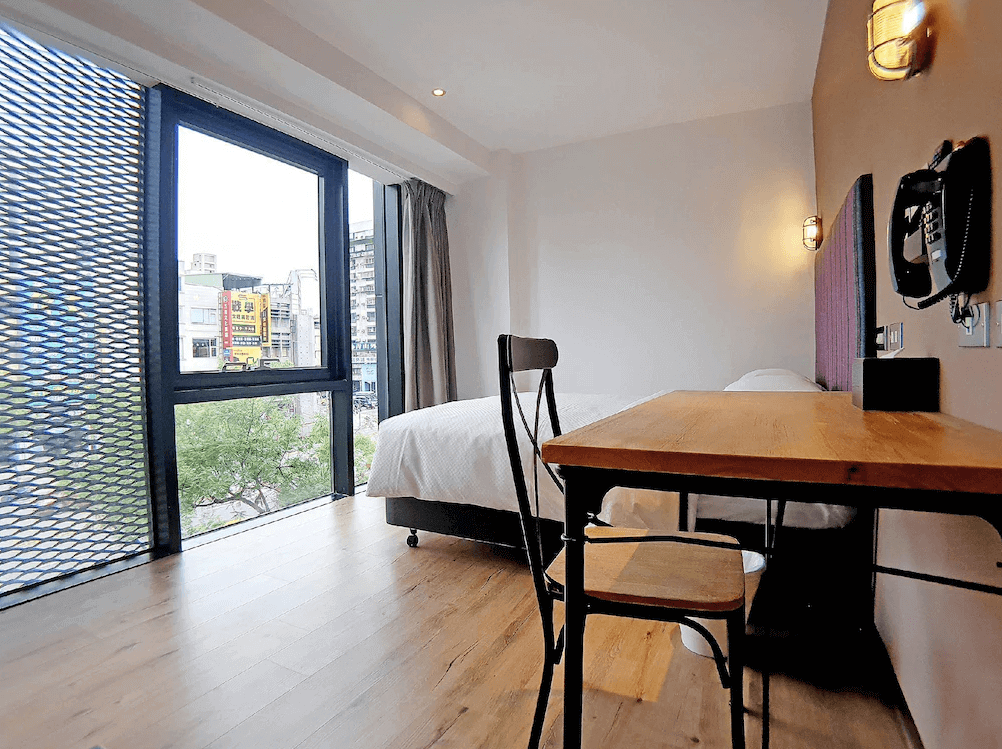 【高雄寵物友善住宿】河堤漫旅 站前館的雙人房，設有大片落地窗可眺望市景，房內配有木質書桌與鐵椅，地板為溫暖色調的木質地板，整體空間清新舒適，採光良好。